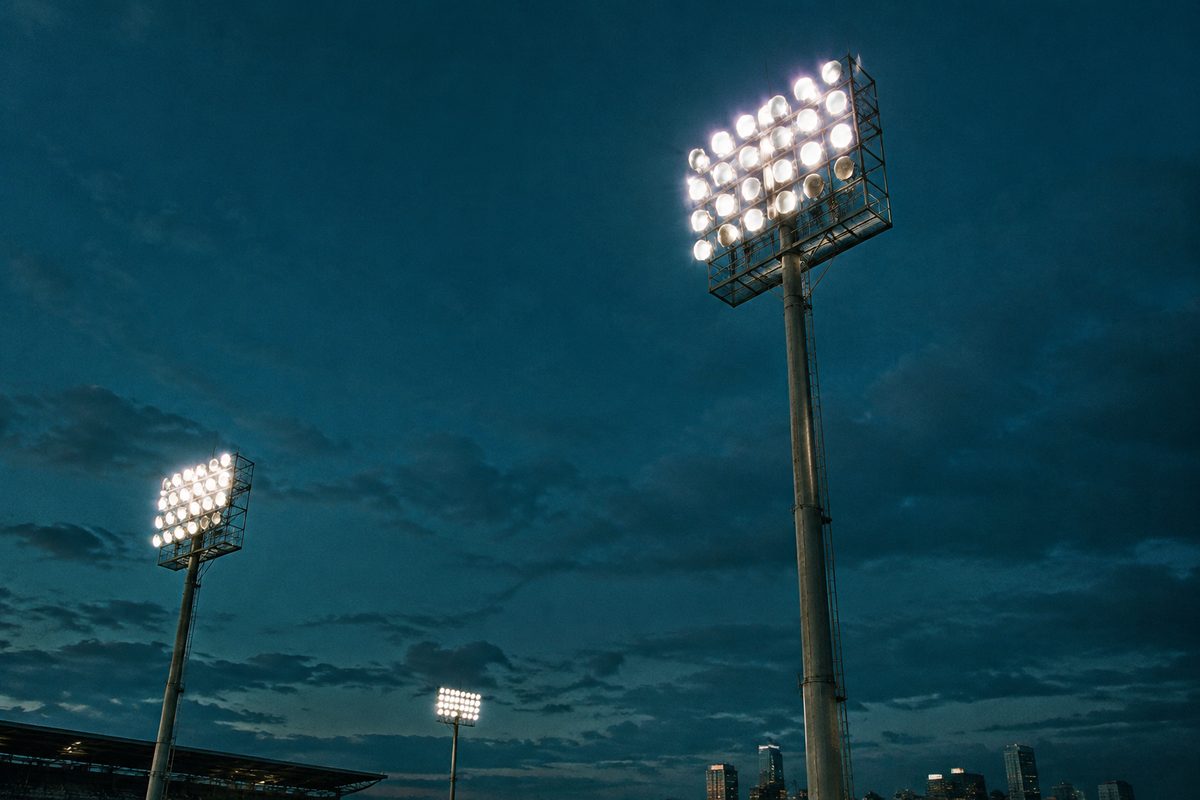 Stadium floodlight towers glowing brightly against a dark teal evening sky.